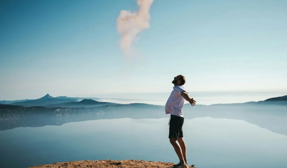 Mężczyzna stojący na klifie nad spokojnym jeziorem z rozłożonymi ramionami, symbol szczęścia i wolności