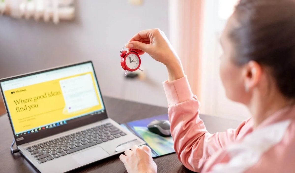 Kobieta pracująca przy laptopie trzymająca w dłoni mały budzik – symbol zarządzania czasem i produktywności